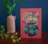 Card with floral design on a brick background, placed on a dark surface with a vase and plant in the background.