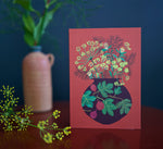 Card with floral design on a brick background, placed on a dark surface with a vase and plant in the background.